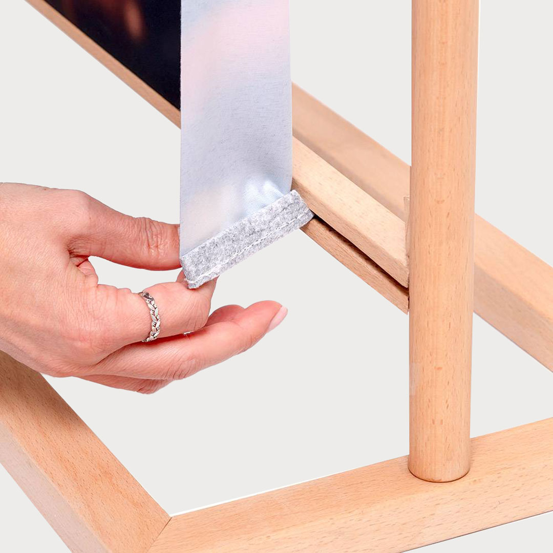 Das untere Ende eines bedruckten Stoffbanners, versehen mit einem grauen Klettverschluss-Saum, wird in die schmale Nut eines hellen Holzrahmens eingeführt.